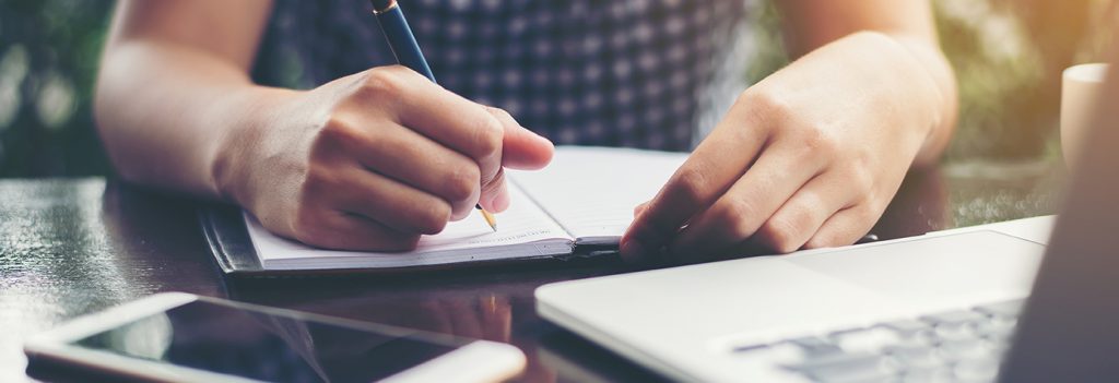 Close up of a woman writing in a notebook in front of a laptop