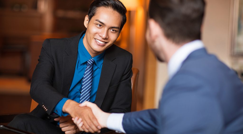 Closeup of two smiling business men shaking hands in cafe. One man is sitting back to camera.