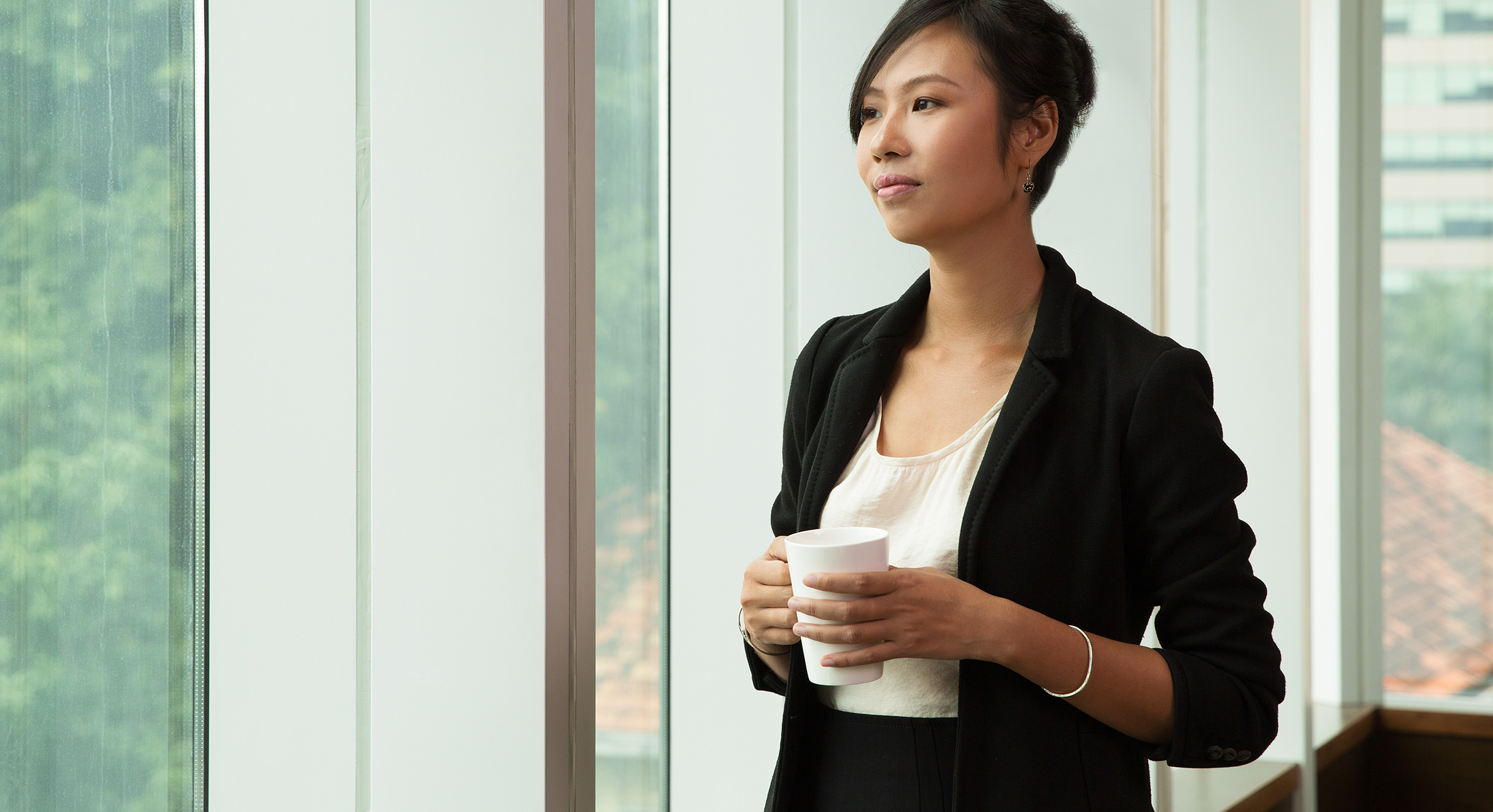 Portrait of pensive Asian businesswoman wearing costume standing in office holding coffee cup and looking out window
