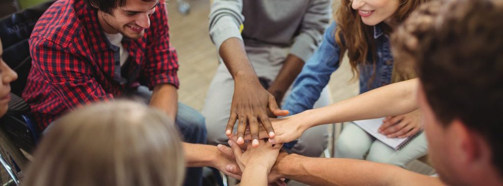 A group of people of differing races and abilities all placing their hand on top of each other in a circle. 