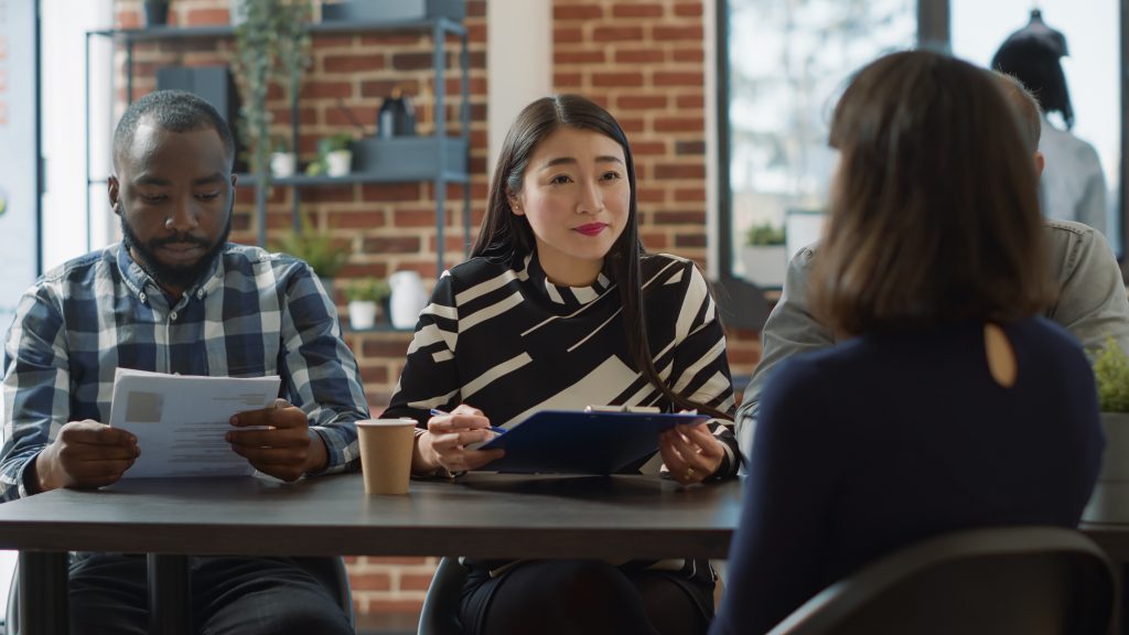 Diverse group of employees talking to applicant at job interview, asking about work experience and business expertise. HR team looking at cv files and interviewing woman at meeting.